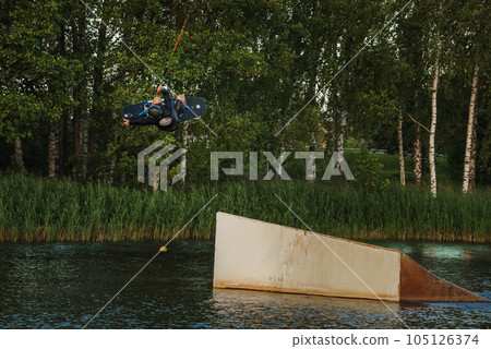 Beautiful view of young man rider holding rope and making extreme jump on wakeboard. Wakeboarding and water sports activity. Low angle shot of man wakeboarding on a lake. Man water skiing at sunset. 105126374