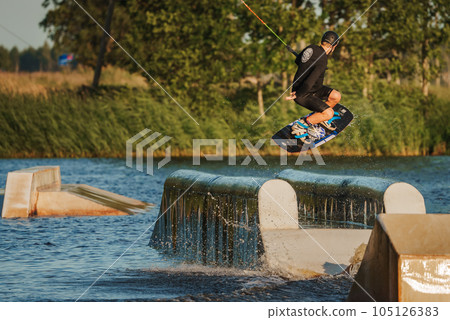 Beautiful view of young man rider holding rope and making extreme jump on wakeboard. Wakeboarding and water sports activity. Low angle shot of man wakeboarding on a lake. Man water skiing at sunset. 105126383