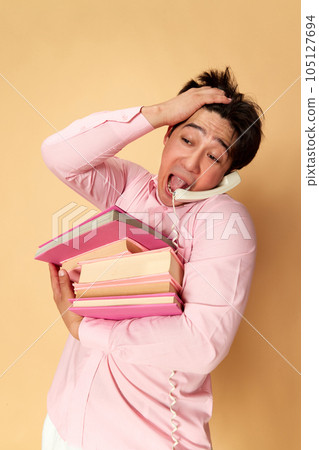 Portrait of young emotional man talking on phone, carrying many books against yellow studio background. Deadlines 105127694