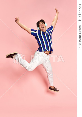 Full-length portrait of young emotional man listening to music in headphones and jumping against pink studio background 105127783