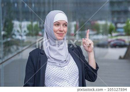 Portrait of business woman in hijab and suit holding index finger up outdoors. Idea. Portrait of business woman in hijab and suit holding index finger up outdoors. Idea. 105127894