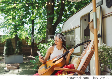 Happy hippie girl is having a good time with playing on guitar with smartphone in her hand in camper trailer. Holiday, vacation, trip concept. 105129499
