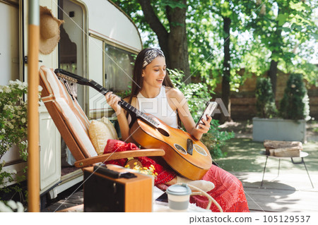 Happy hippie girl is having a good time with playing on guitar with smartphone in her hand in camper trailer. Holiday, vacation, trip concept. 105129537