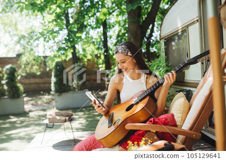 Happy hippie girl is having a good time with playing on guitar with smartphone in her hand in camper trailer. Holiday, vacation, trip concept. 105129641
