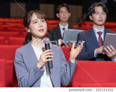 A young woman asking a question with a microphone in her hand while attending a seminar in a hall 105131422