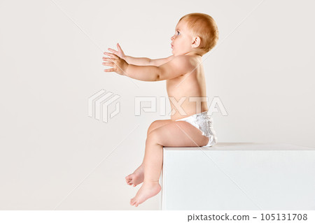 Portrait of little beautiful baby, toddler in diaper calmly sitting, looking and playing against white studio background 105131708