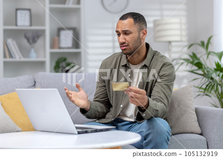 Financial problems. Upset African American young man sitting at home in front of laptop, looking worriedly at screen, holding credit card and spreading hands. Financial problems. Upset African American young man sitting at home in front of laptop, looking worriedly at screen, holding credit card and spreading hands. 105132919