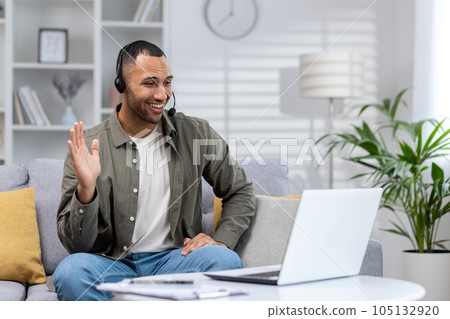 A young African American man wearing a headset conducts an online business video call training, meeting and conference with the team from home. Says hello and waves to the camera. 105132920