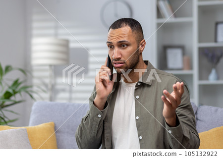 Portrait of upset young hispanic man talking on the phone at home. Worriedly spreads his hands, solves problems on the phone, received unpleasant news. Portrait of upset young hispanic man talking on the phone at home. Worriedly spreads his hands, solves problems on the phone, received unpleasant news. 105132922