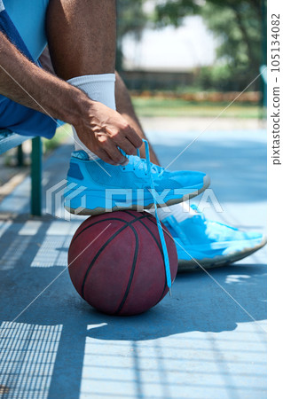 Close-up image of male legs in sportswear standing on basketball ball on outdoor sportsground. Man tying shoelaces 105134082