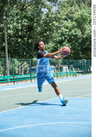Full-length image of competitive young man in blue uniform playing basketball, throwing ball. Outdoor sportsground Full-length image of competitive young man in blue uniform playing basketball, throwing ball. Outdoor sportsground 105134090