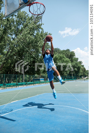 Full-length dynamic image of young guy in blue uniform jumping, playing basketball outdoors on sportsground on warm sunny day Full-length dynamic image of young guy in blue uniform jumping, playing basketball outdoors on sportsground on warm sunny day 105134091