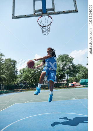 Full-length image of competitive young man in blue uniform playing basketball, throwing ball. Outdoor sportsground 105134092