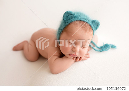 A cute newborn boy in the first days of life sleeps naked against a background of white fabric. Blue azure cap with ears like a bear. A cute newborn boy in the first days of life sleeps naked against a background of white fabric. Blue azure cap with ears like a bear. 105136072