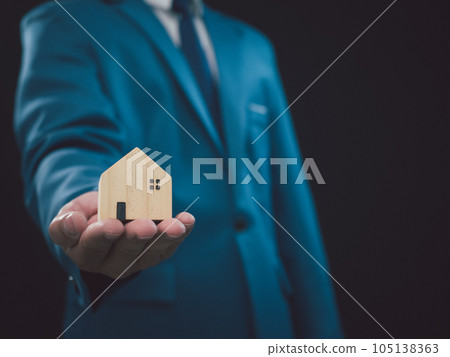 Concept of selling a house. Businessman is holding a model house on a dark background. Real estate agent offer house, property insurance and security, affordable housing concepts, home insurance 105138363