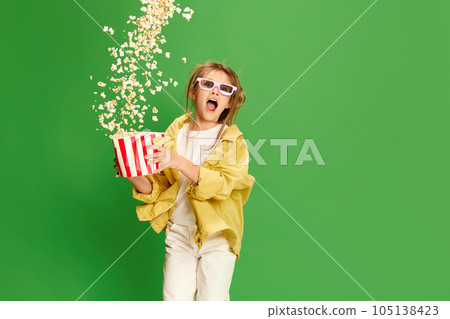 Happy excited little girl in 3d glasses standing with pop corn backet and shouting with delight over green background. 105138423