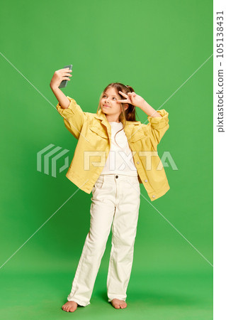 Full-length shot of little girl dressed stylishly taking selfie on cell phone and showing v-sign isolated on green background. 105138431
