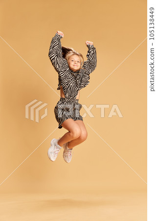 Full size portrait of beautiful blonde child in cute animal print suit jumping and looking at camera over sandy background 105138449
