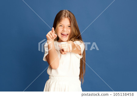 Portrait of little smiling child looking and pointing her finger at camera on blue background. Concept happy mood, beauty, fashion 105138458