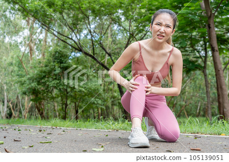 Young atractive Asian woman with knee pain putting her hands over her troubled right knee while sitting down at running track of a local park 105139051