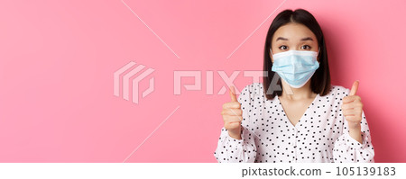 Covid-19, pandemic and lifestyle concept. Supportive asian female in face mask showing thumbs-up, approve and praise good work, standing over pink background Covid-19, pandemic and lifestyle concept. Supportive asian female in face mask showing thumbs-up, approve and praise good work, standing over pink background 105139183