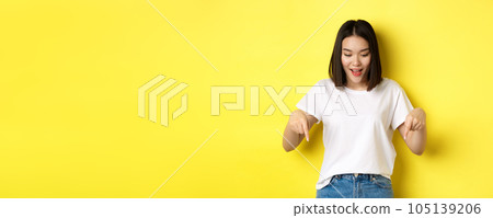 Beauty and fashion concept. Beautiful asian woman in white t-shirt pointing fingers down, demonstrate logo standing over yellow background Beauty and fashion concept. Beautiful asian woman in white t-shirt pointing fingers down, demonstrate logo standing over yellow background 105139206