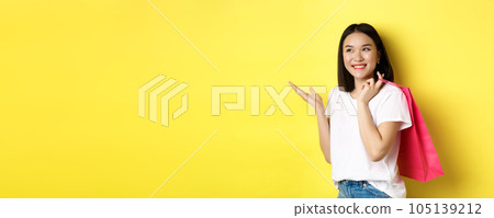 Cheerful asian female shopper looking amused, holding shopping bag and pointing hand left at store banner, standing over yellow background 105139212