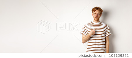 Confused redhead man in glasses pointing at himself, looking questioned at camera, white background 105139221