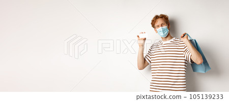 Concept of covid-19 and lifestyle. Cheerful young man with red hair, wear medical mask, showing shopping bag from store and plastic credit card 105139233