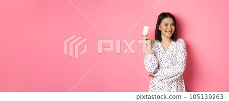 Shopping concept. Confident and happy asian woman holding credit card and smiling satisfied, standing against pink background Shopping concept. Confident and happy asian woman holding credit card and smiling satisfied, standing against pink background 105139263