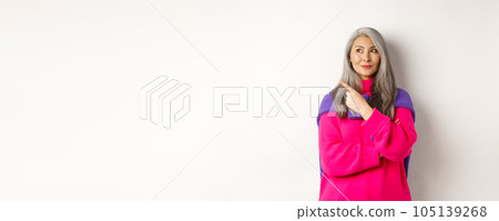 Stylish modern asian elderly woman pointing at upper left corner, looking at logo with pleased smile, standing in pink sweater over white background 105139268