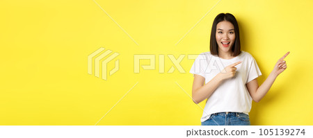 Beauty and fashion concept. Beautiful asian woman in white t-shirt pointing fingers right, demonstrate logo standing over yellow background Beauty and fashion concept. Beautiful asian woman in white t-shirt pointing fingers right, demonstrate logo standing over yellow background 105139274