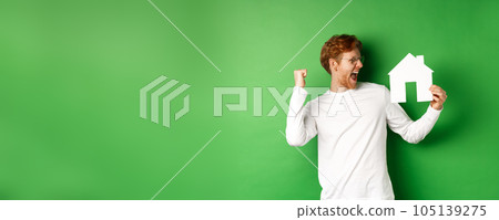 Real estate. Cheerful redhead man in glasses celebrating, buying a house, showing paper home cutout and scream with joy, standing over green background Real estate. Cheerful redhead man in glasses celebrating, buying a house, showing paper home cutout and scream with joy, standing over green background 105139275