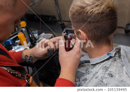 The hands of a guy, a cool hairdresser, a professional hold a machine and a hair brush against the background of a client's haircut in the salon The hands of a guy, a cool hairdresser, a professional hold a machine and a hair brush against the background of a client's haircut in the salon 105139495