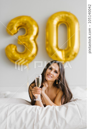 Happy young woman meets her thirtieth birthday in bed, holding a glass of champagne in her hands and smiling 105140042