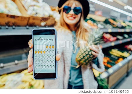 Close up phone with online mobile app of Veganuary diet calendar. Stylish fashion woman holding pineapple in the supermarket store during selecting products. Healthy eating, go vegan. Selective focus Close up phone with online mobile app of Veganuary diet calendar. Stylish fashion woman holding pineapple in the supermarket store during selecting products. Healthy eating, go vegan. Selective focus 105140465