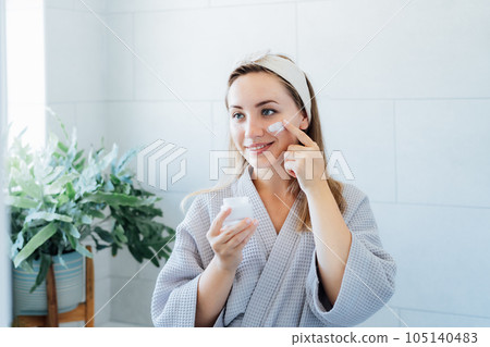 Smiling young woman with skin problems in bathrobe takes care face skin at home. She looking in the mirror and aplying cream on the face in bathroom. Acne skin care every day treatment process. 105140483