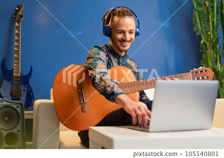Young man sitting in his room and watching guitar tutorial on laptop 105140801