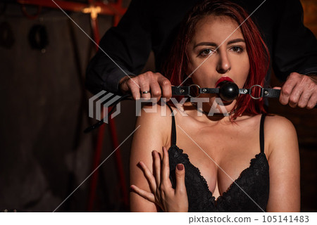 A man dominates and puts a gag in his partner mouth. BDSM concept. Portrait of a woman in seductive underwear with an intimate toy in her mouth. Sexy couple plays love games. A man dominates and puts a gag in his partner mouth. BDSM concept. Portrait of a woman in seductive underwear with an intimate toy in her mouth. Sexy couple plays love games. 105141483