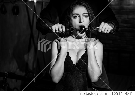 A man dominates and puts a gag in his partner mouth. BDSM concept. Portrait of a woman in seductive underwear with an intimate toy in her mouth. Sexy couple plays love games. A man dominates and puts a gag in his partner mouth. BDSM concept. Portrait of a woman in seductive underwear with an intimate toy in her mouth. Sexy couple plays love games. 105141487