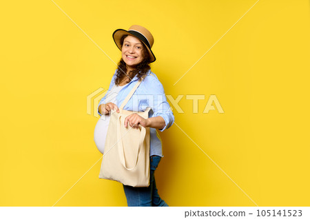 Beautiful pregnant woman smiles cutely looking at camera, carrying eco linen shopping bag on isolated yellow background Beautiful pregnant woman smiles cutely looking at camera, carrying eco linen shopping bag on isolated yellow background 105141523