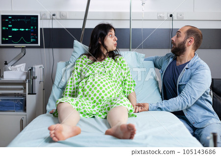 Woman with pregnancy lying in hospital ward bed with husband beside her waiting for child delivery. Patient preparing for labor having pain contractions, asking for doctor to come and start surgery 105143686