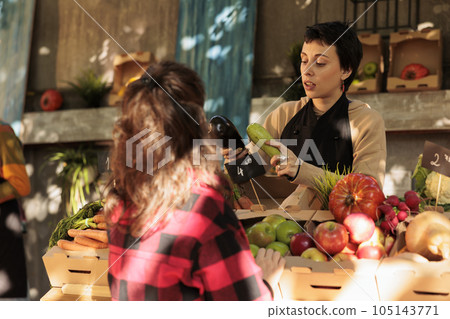 Young female farmer talking with customer while selling fresh locally grown vegetables at farmers market, local vendor helping consumer to choose veggies, explaining health benefits of organic food Young female farmer talking with customer while selling fresh locally grown vegetables at farmers market, local vendor helping consumer to choose veggies, explaining health benefits of organic food 105143771