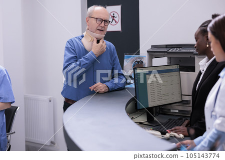 Injured elderly patient with cervical neck collar standing at hospital registration counter discussing about appointment with receptionist. Man with medical brace waiting to meet with traumatologist. Injured elderly patient with cervical neck collar standing at hospital registration counter discussing about appointment with receptionist. Man with medical brace waiting to meet with traumatologist. 105143774
