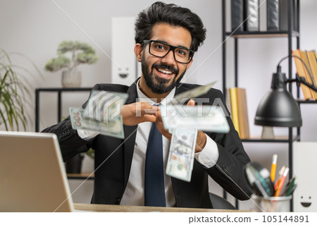 Cheerful rich business man working on office laptop wasting throwing money to camera profit win 105144891