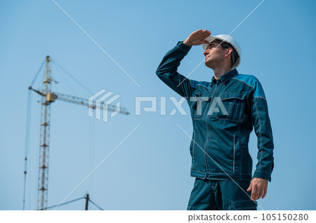 A builder in work clothes and a helmet stands on a construction site against the background of a construction crane.  105150280