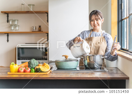 圍裙的亞裔婦女在廚房的電水壺裡放熱水 圍裙的亞裔婦女在廚房的電水壺裡放熱水 105151450