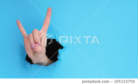 Horns sign. Rock cool. Female fan hand showing fingers up inside breakthrough paper hole on blue ripped wall background with empty space. 105153750