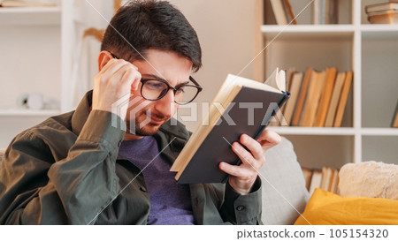 Book plot. Hard literature. Poor eyesight. Tensed frowning man in glasses struggling with complicated confusing paper novel on couch at home. 105154320