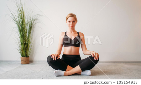 Indoor yoga. Home training. Health wellbeing. Confident relaxed woman sitting cross-legged in lotus position on floor at light studio interior with empty space. Indoor yoga. Home training. Health wellbeing. Confident relaxed woman sitting cross-legged in lotus position on floor at light studio interior with empty space. 105154915